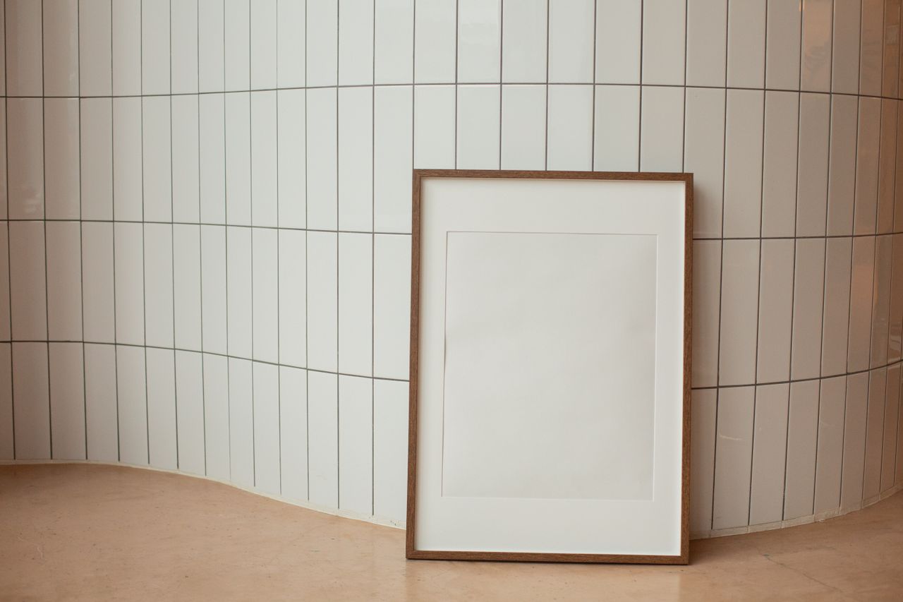 Empty white wooden photo frame placed on floor against curvy tiled wall in light room
